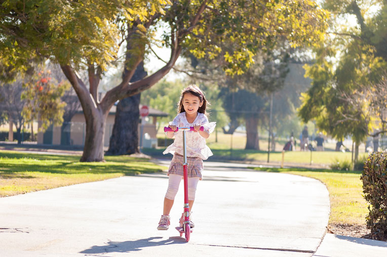 Child riding a scooter on a wide path surrounded by greenery and trees